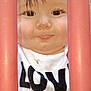 child, toddler, face, closeup, curious, red_bars, white_shirt, text_on_clothing, outdoor, young_child, smile, eyes, hair, expression, portrait, peeking, playground, bars, innocence, baby