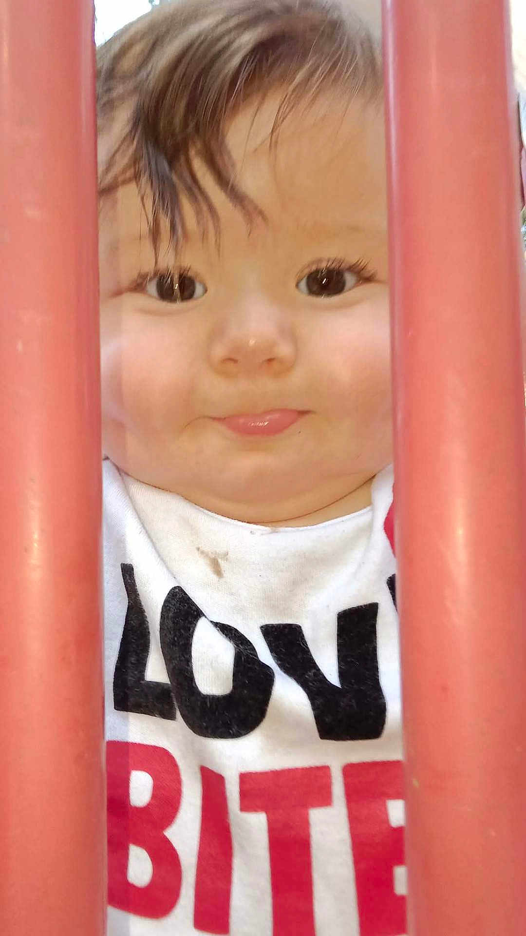 Caspain joined the competition — help win amazing prizes! child, toddler, face, closeup, curious, red_bars, white_shirt, text_on_clothing, outdoor, young_child, smile, eyes, hair, expression, portrait, peeking, playground, bars, innocence, baby