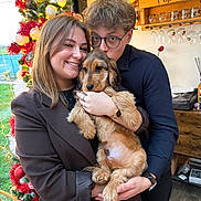 Arès participe au concours pour gagner de l'argent avec cette photo : christmas_tree, holiday_decor, couple, woman, man, puppy, dog, glasses, smile, indoor, festive, warm_lighting, hug, red_decorations, greenery, watch, wood_floor, window, furniture, celebration