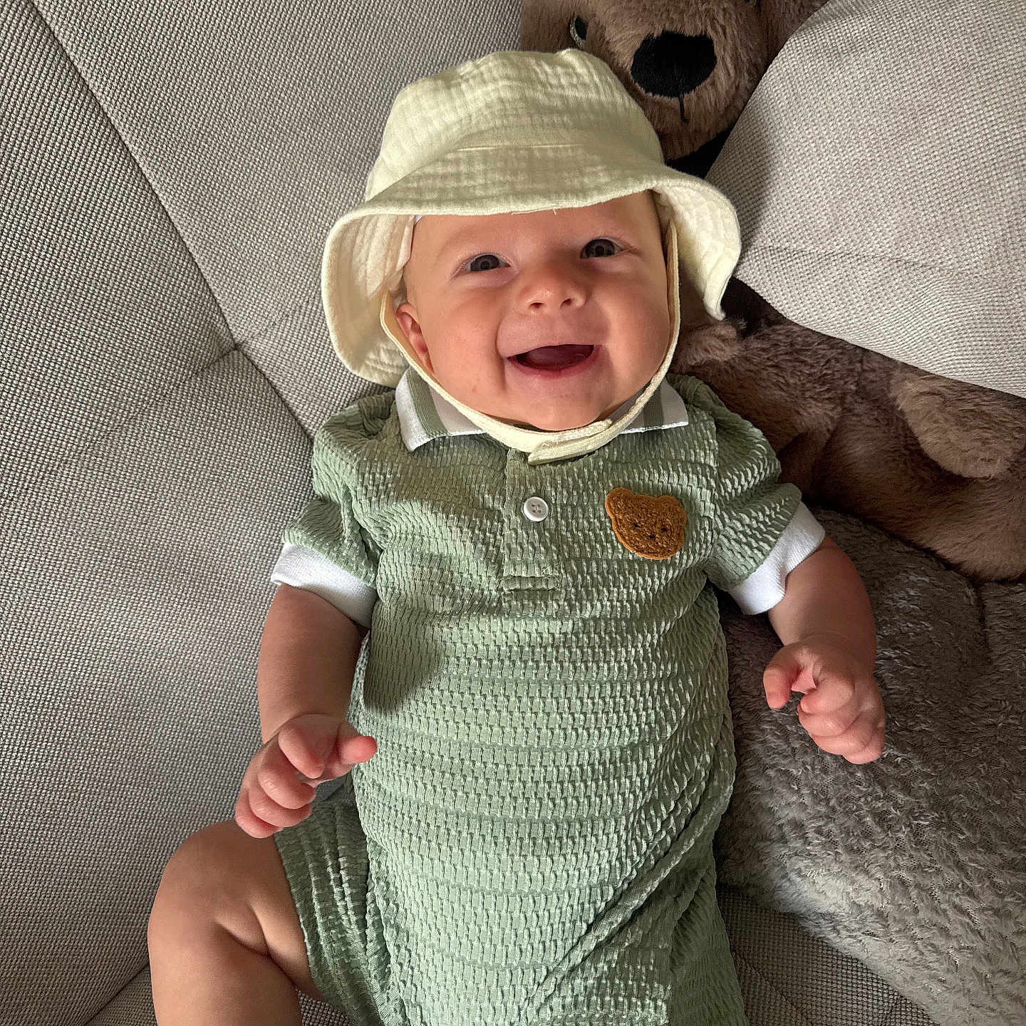 Benny is registered to the contest to win money with this photo: baby, infant, smiling, hat, plush_toy, kangaroo, couch, textured_fabric, green_outfit, child, happy, indoor, cute, toddler, soft_toy, seated, portrait, cozy, playful, relaxed