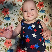 Paisley is registered to the contest to win money with this photo: baby, child, smiling, face, hand, tank_top, stars_pattern, shorts, red_checkered, floral_background, blanket, lying_down, cute, happy, infant, person, portrait, indoors, colorful, playful