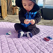 Matéo a rejoint le concours — aidez-le/la à gagner de superbes lots ! baby, toddler, child, sitting, hoodie, hooded_jacket, toy_cars, pacifier, blanket, purple_blanket, socks, outdoor, patio, paved_floor, surprised_expression, hands, face, playtime, daylight, garden