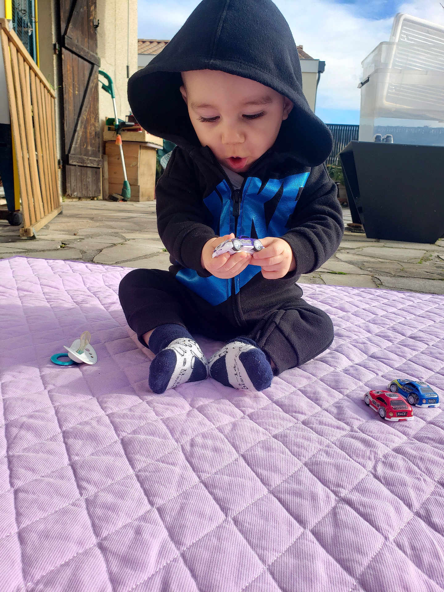 Matéo a rejoint le concours — aidez-le/la à gagner de superbes lots ! baby, toddler, child, sitting, hoodie, hooded_jacket, toy_cars, pacifier, blanket, purple_blanket, socks, outdoor, patio, paved_floor, surprised_expression, hands, face, playtime, daylight, garden