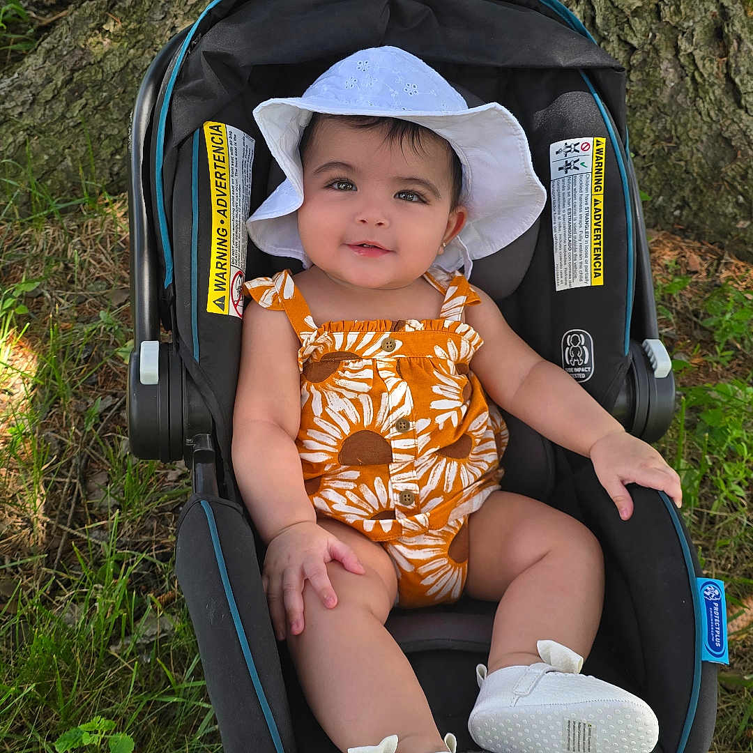 Julia joined the competition — help win amazing prizes! baby, baby_shoes, car_seat, child, cute, footwear, grass, greenery, happy, infant, nature, outdoor, portrait, romper, seat, smiling, summer_clothing, sunlight, tree_trunk, white_hat