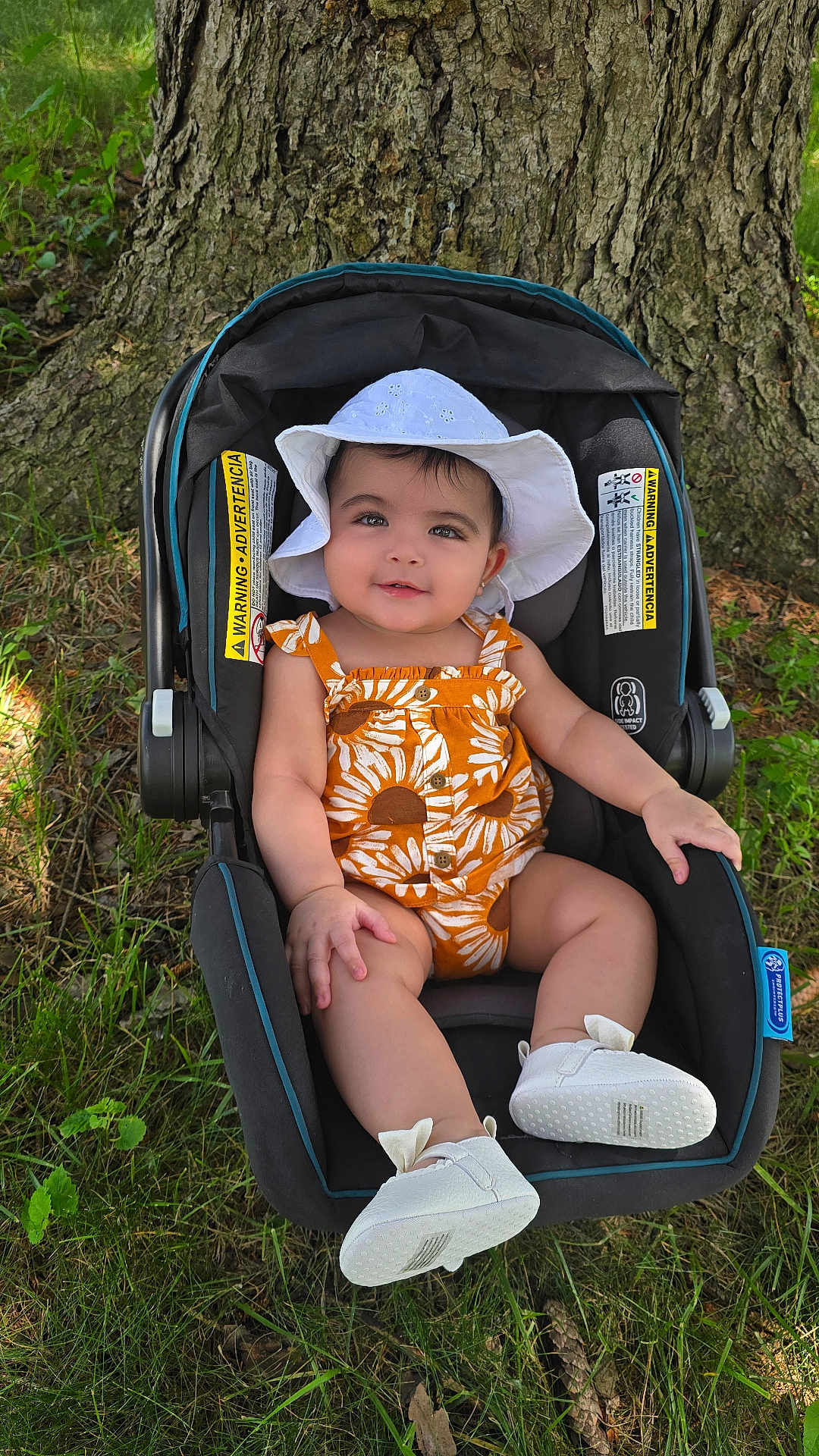 Julia joined the competition — help win amazing prizes! baby, child, car_seat, white_hat, romper, outdoor, tree_trunk, grass, sunlight, smiling, footwear, infant, nature, greenery, cute, summer_clothing, seat, baby_shoes, portrait, happy