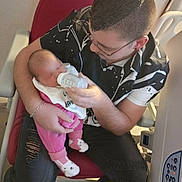 Gabriela participe au concours pour gagner de l'argent avec cette photo : baby, adult, feeding, bottle, chair, pink_clothing, black_shirt, ripped_jeans, socks, hospital_bed, medical_equipment, glasses, bracelet, indoor, person, seated, floor, love, care, tenderness
