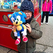 Mathéo participe au concours pour gagner de l'argent avec cette photo : child, plush_toy, sonic_the_hedgehog, winter_clothing, hat, glasses, claw_machine, arcade, outdoor, person, toy, smile, jacket, woman, pink_jacket, concrete, game, happy, crowd, casual