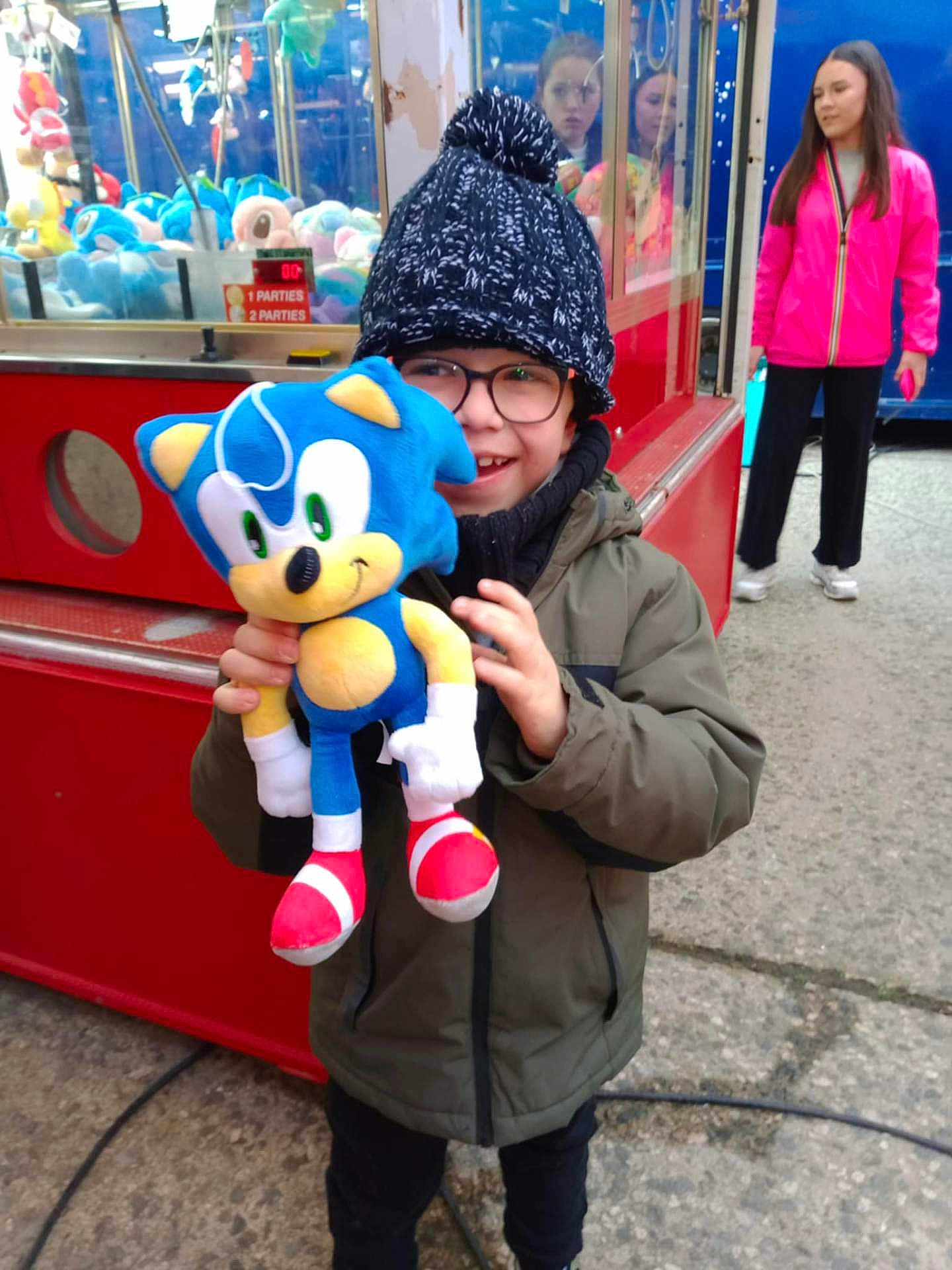 Mathéo participe au concours pour gagner de l'argent avec cette photo : child, plush_toy, sonic_the_hedgehog, winter_clothing, hat, glasses, claw_machine, arcade, outdoor, person, toy, smile, jacket, woman, pink_jacket, concrete, game, happy, crowd, casual