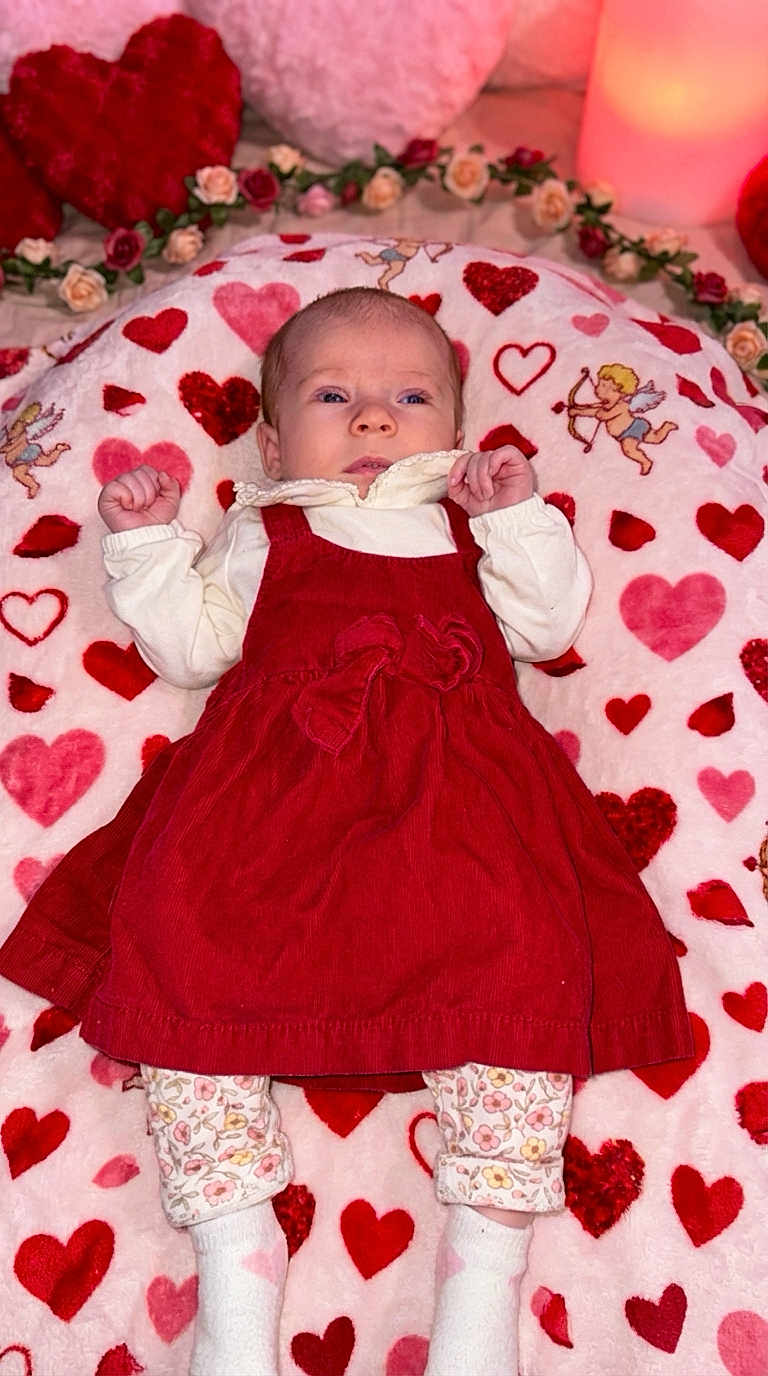 Madeline joined the competition — help win amazing prizes! baby, infant, child, red_dress, heart_blanket, valentine_theme, blanket, cupid_illustration, pillow, floral_pattern, socks, lying_down, portrait, cute, decorations, rose_garland, plush, cozy, candle, neutral_expression
