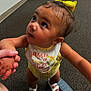 baby, bib, carpet, child, clothing, curious, cute, expression, footwear, hair, holding_hands, indoor, looking_up, person, skin, standing, toddler, white_sandals, yellow_bow, young_child