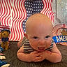 american_flag, baby, background, celebration, child, cute, decorations, face, festive, happy, holiday, indoor, patriotic, portrait, red_white_blue, smiling, stars, striped_clothing, toy, tummy_pose
