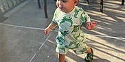 Jonah is registered to the contest to win money with this photo: toddler, child, outdoor, sunlight, shadow, concrete_floor, green_clothing, patterned_outfit, shoes, patio, table, chair, railing, daylight, person, walking, shorts, cute, baby, happy