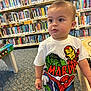 Jonah is registered to the contest to win money with this photo: toddler, child, marvel, superhero, library, books, bookshelf, carpet, clothing, standing, indoor, person, curious, short_hair, young_child, casual_wear, playful, portrait, daylight, hand_on_table