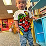 Jonah is registered to the contest to win money with this photo: toddler, child, indoor, playroom, toy, corn, marvel, tshirt, jeans, shoes, bookshelf, carpet, furniture, person, young_child, colorful, casual_clothing, standing, expression, play