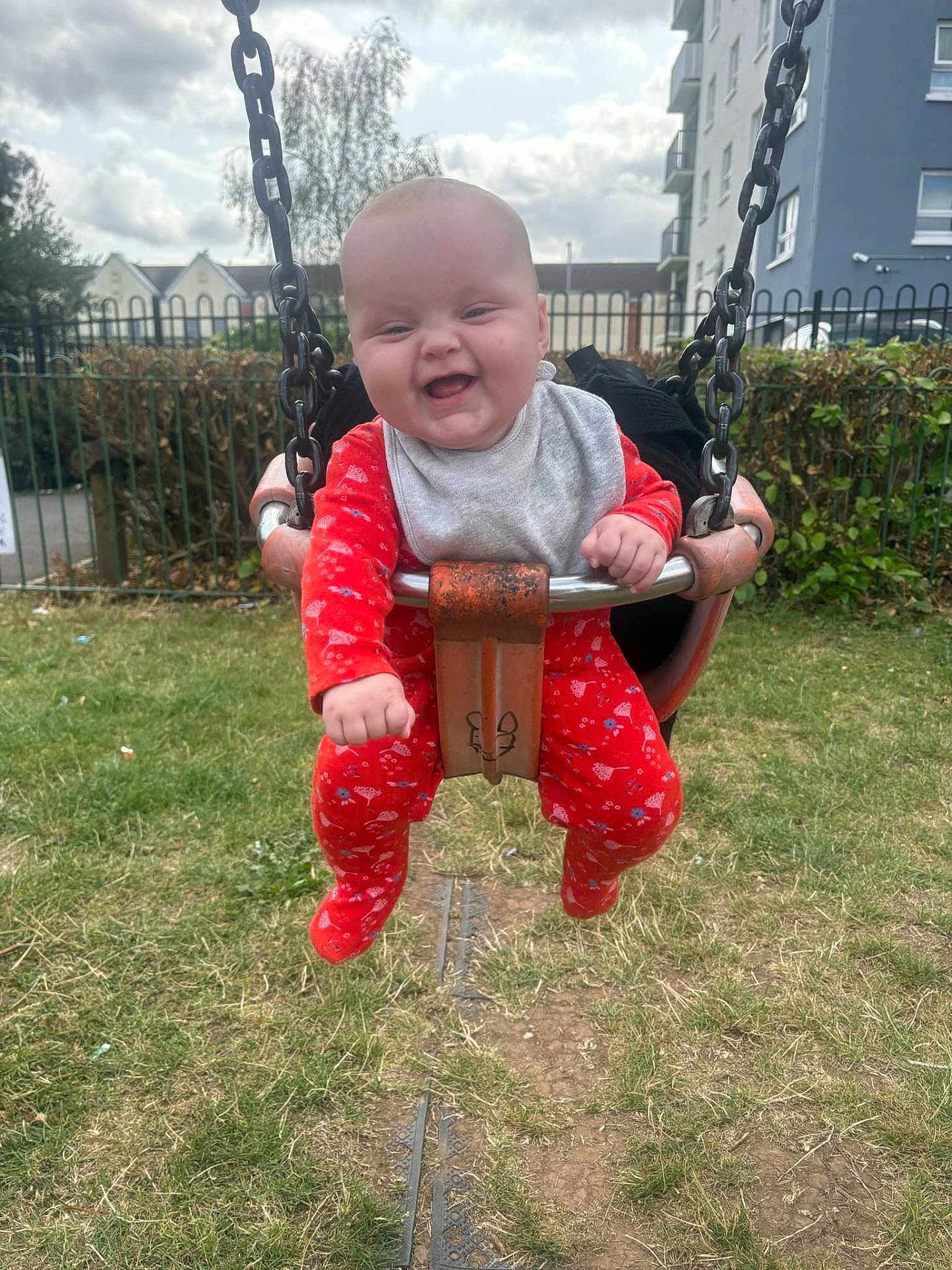 Luna joined the competition — help win amazing prizes! baby, swing, playground, outdoor, grass, fence, smiling, child, red_clothing, onesie, chain, park, happy, face, person, daytime, building, sky, fun, joy