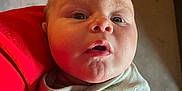 Samuel is registered to the contest to win money with this photo: baby, infant, wrapped, blanket, face, expression, wide_eyes, mouth_open, red_clothing, person_holding, indoor, closeup, skin, head, child, newborn, soft_light, curious, portrait, candid