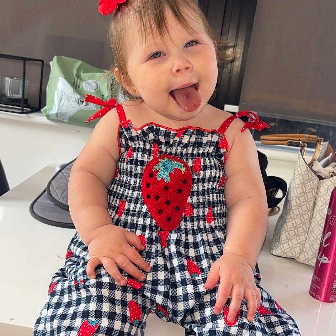 Tilly-May is registered to the contest to win money with this photo: baby, bag, can, checkered_clothing, child, cute, funny_face, happy, indoor, person, pigtails, playful, portrait, red_bows, sitting, smiling, strawberry_pattern, table, toddler, tongue_out