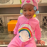 Khalyla joined the competition — help win amazing prizes! toddler, child, pink_onesie, rainbow, hat, flower_clips, smiling, indoor, glass_table, fan, orange_box, happy, cute, sitting, person, young_child, cozy, colorful, playful, portrait