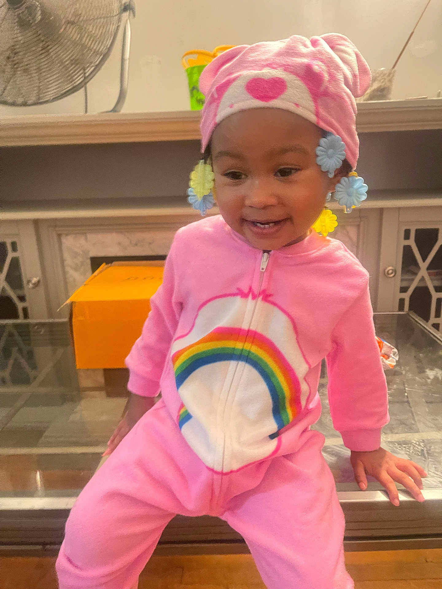 Khalyla joined the competition — help win amazing prizes! toddler, child, pink_onesie, rainbow, hat, flower_clips, smiling, indoor, glass_table, fan, orange_box, happy, cute, sitting, person, young_child, cozy, colorful, playful, portrait
