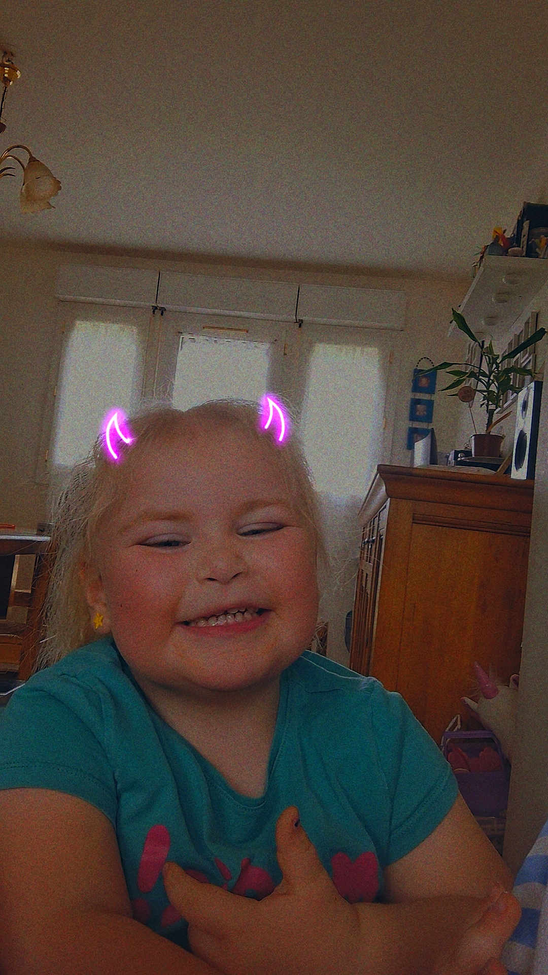 Lynaë participe au concours pour gagner de l'argent avec cette photo : blonde_hair, blue_shirt, cabinet, child, crossed_arms, curtains, devil_horns_filter, earrings, furniture, girl, happy, home, indoor, plant, playful, portrait, smiling, toy, window, yellow_earrings