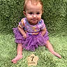 Julie joined the competition — help win amazing prizes! baby, child, purple_tutu, colorful_shirt, sitting, green_rug, fluffy_texture, milestone_marker, wooden_sign, 7_months, portrait, cute, infant, smiling, indoor, floor, feet, hands, looking_at_camera, close_up