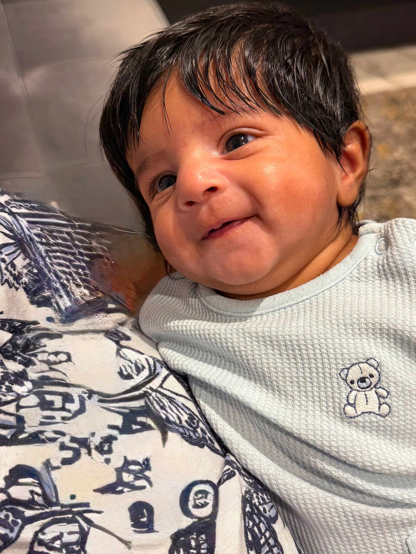 Issac is registered to the contest to win money with this photo: baby, infant, child, smile, face, black_hair, blue_eyes, white_clothing, teddy_bear, patterned_fabric, close_up, cute, portrait, indoors, soft_lighting, happy, young, person, head, skin