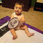 Kailee is registered to the contest to win money with this photo: baby, barefoot, blanket, brown_hair, carpet, child, close_up, cozy, family_photo, happy, home, indoors, infant, milestone_sign, onesie, portrait, quilt, sitting, smiling, wooden_furniture