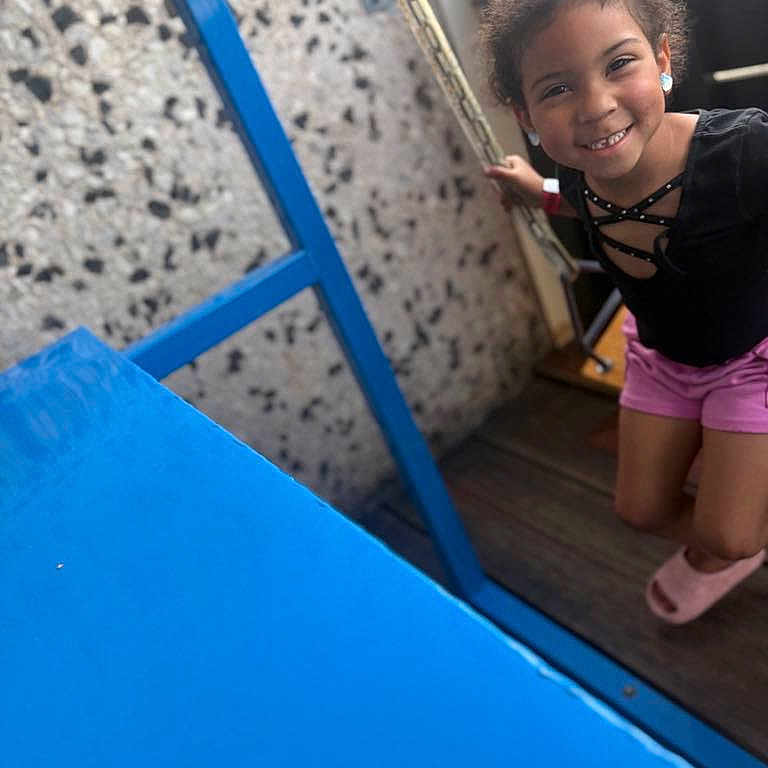 Sevyn is registered to the contest to win money with this photo: black_top, blue_frame, candid, casual, child, curly_hair, earrings, girl, happy, holding, indoor, joyful, person, pink_shorts, play, portrait, smiling, swing, wall, wooden_floor