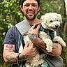 Bud is registered to the contest to win money with this photo: man, dog, pet, tattoo, smile, outdoor, forest, nature, cap, tshirt, white_dog, curly_fur, leash, harness, happy, portrait, holding, arm_tattoo, greenery, daylight