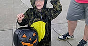 Jasper joined the competition — help win amazing prizes! adult, black, casual_clothing, child, concrete, costume, dinosaur_costume, festive, green, halloween, orange, outdoor, person, pumpkin_bucket, shoes, sidewalk, smiling, street, trick_or_treat, yellow