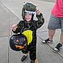 child, dinosaur_costume, halloween, pumpkin_bucket, sidewalk, outdoor, smiling, costume, trick_or_treat, person, shoes, street, concrete, adult, casual_clothing, green, yellow, black, orange, festive