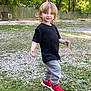 toddler, child, outdoor, grass, gravel, tree, fence, sneakers, red_shoes, black_tshirt, gray_pants, playful, smiling, person, nature, daylight, young_child, curly_hair, walking, casual_clothing