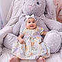 baby, child, plush_toy, stuffed_animal, bunny, dress, headband, smiling, sitting, indoor, cozy, cute, soft_toy, happy, infant, decor, bed, comfort, person, portrait