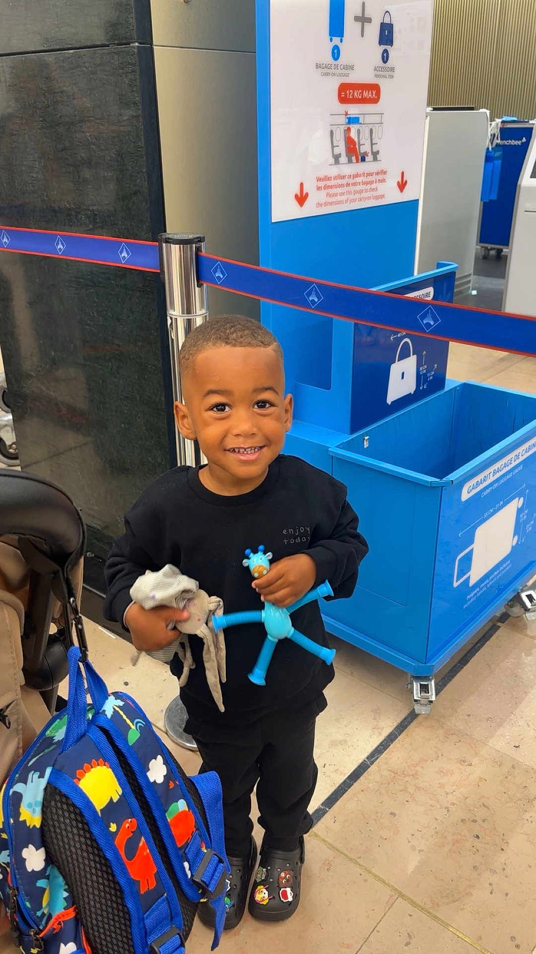 Nahïl a rejoint le concours — aidez-le/la à gagner de superbes lots ! child, smiling, toy, backpack, airport, luggage, blue, black_clothing, floor, sign, person, happy, indoor, shoes, travel, holding, young_child, dinosaur_pattern, waiting_area, luggage_bin