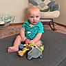 baby, child, indoor, toy, plush_toy, carpet, rocking_chair, quilted_mat, green_shirt, dinosaur_pattern, curious_expression, bald_head, sitting, playful, nursery, baby_bouncer, furniture, floor, colorful, person