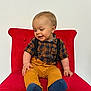 toddler, child, red_chair, plaid_shirt, suspenders, mustard_pants, blue_socks, smiling, sitting, indoor, happy, cute, baby, person, clothing, furniture, portrait, young_child, casual, light_background