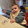 baby, child, high_chair, table, bib, hoodie, indoor, restaurant, wooden_floor, red_chairs, person, face, hand, curious, seated, clothing, cute, infant, expression, casual