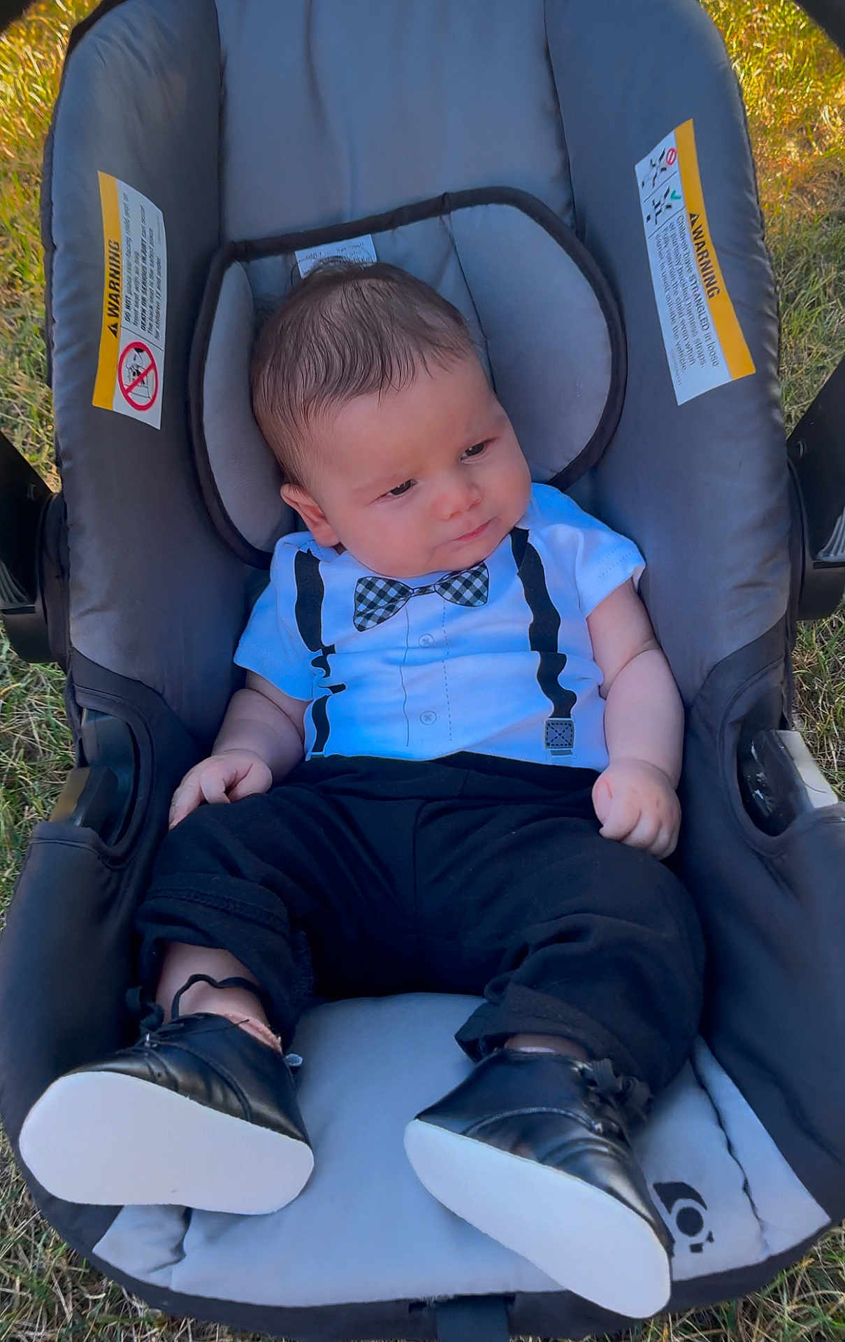 Ahmed Adam is registered to the contest to win money with this photo: baby, infant, car_seat, outdoor, grass, clothing, black_pants, bow_tie, onesie, shoes, footwear, person, young_child, cute, portrait, sitting, sunlight, expression, head, face