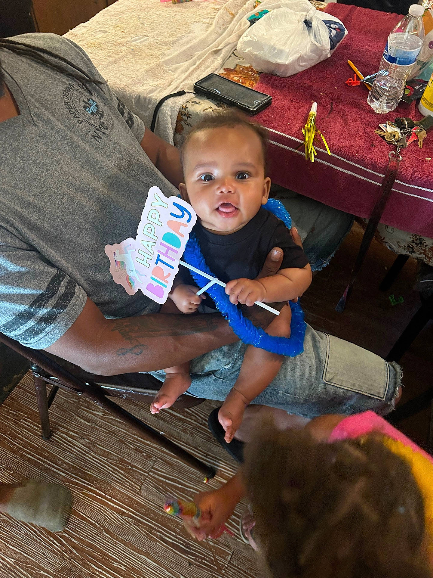 adult, baby, birthday, blue_lei, celebration, child, clothing, family, flooring, hand, happy_birthday_sign, holding, indoor, keys, party, person, sitting, table, toy, water_bottle