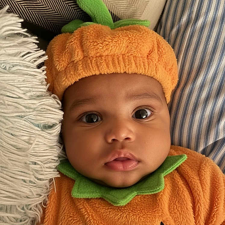 Kacen is registered to the contest to win money with this photo: baby, child, closeup, costume, cute, eyes, fabric, face, green, halloween, hat, holiday, infant, lying_down, orange, pillow, portrait, pumpkin_costume, soft, texture