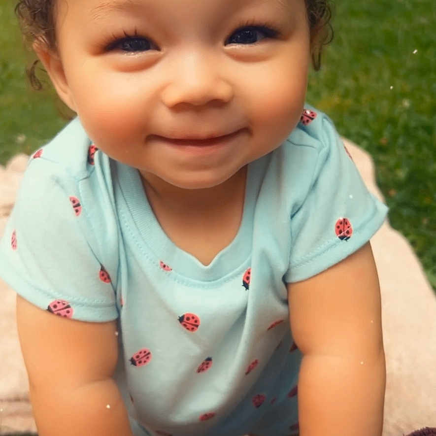 Alani is registered to the contest to win money with this photo: toddler, child, baby, smiling, curly_hair, portrait, outdoor, grass, ladybug, clothing, onesie, heart, sticker, blanket, hands, arms, face, sitting, happy, close_up