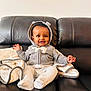 baby, child, smile, hoodie, gray_clothing, blanket, couch, leather_furniture, sitting, happy, infant, indoor, cute, portrait, warm, cozy, hands, feet, headwear, comfortable