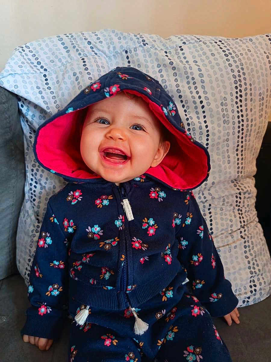 Leyna participe au concours pour gagner de l'argent avec cette photo : baby, cheerful, child, clothing, couch, cute, face, floral_pattern, happy, hoodie, indoor, infant, navy_blue, person, pillow, pink, portrait, seated, smiling, young