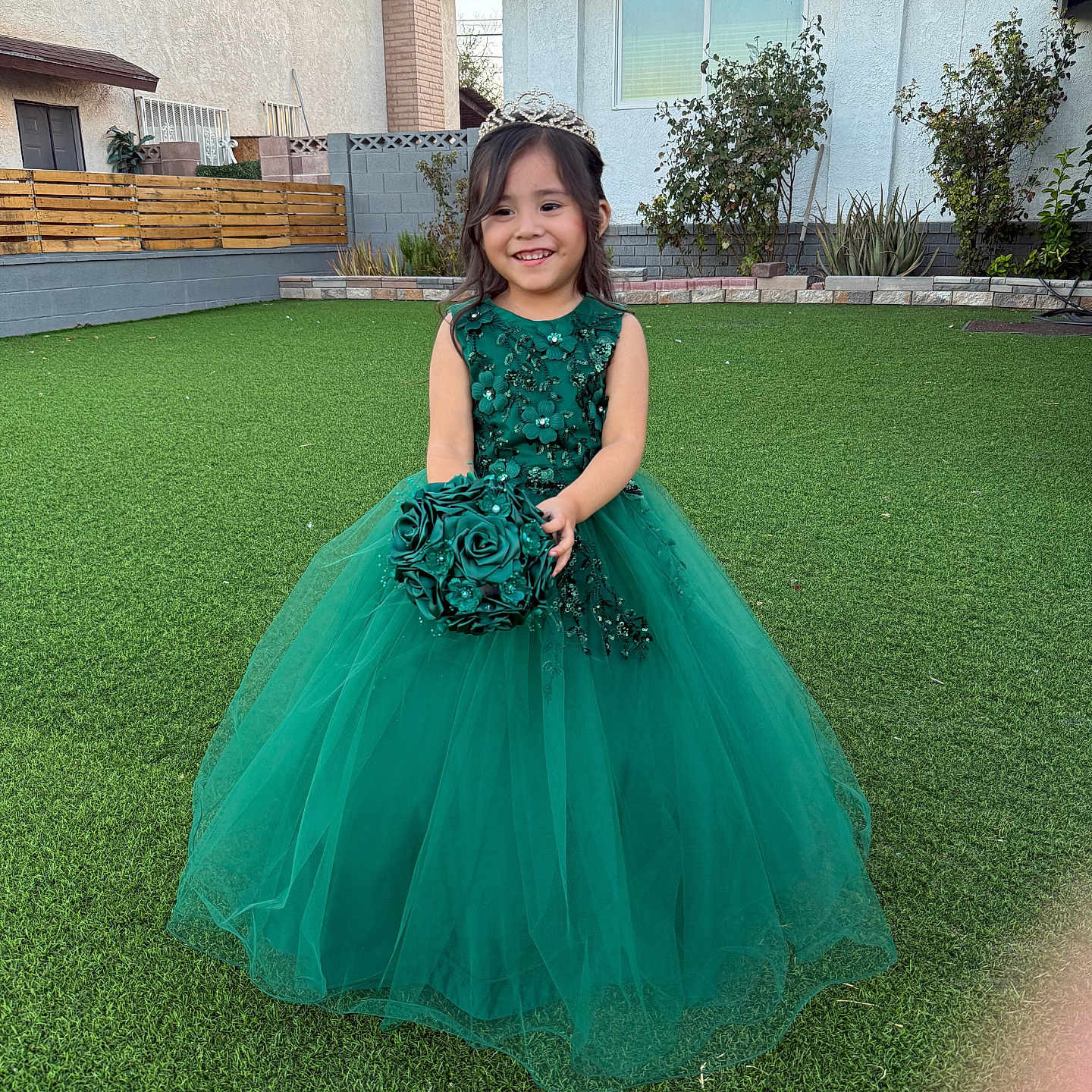 Ledif is registered to the contest to win money with this photo: child, girl, smiling, portrait, outdoor, backyard, garden, green_dress, ball_gown, tiara, formalwear, bouquet, flowers, artificial_grass, house, fence, plants, royal, standing, happy