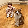Shelby joined the competition — help win amazing prizes! child, toddler, baby, cat, pet, orange_cat, white_cat, sitting, indoor, kitchen, wooden_floor, cabinets, portrait, looking_at_camera, floral_clothing, pajamas, socks, cute, companion, home