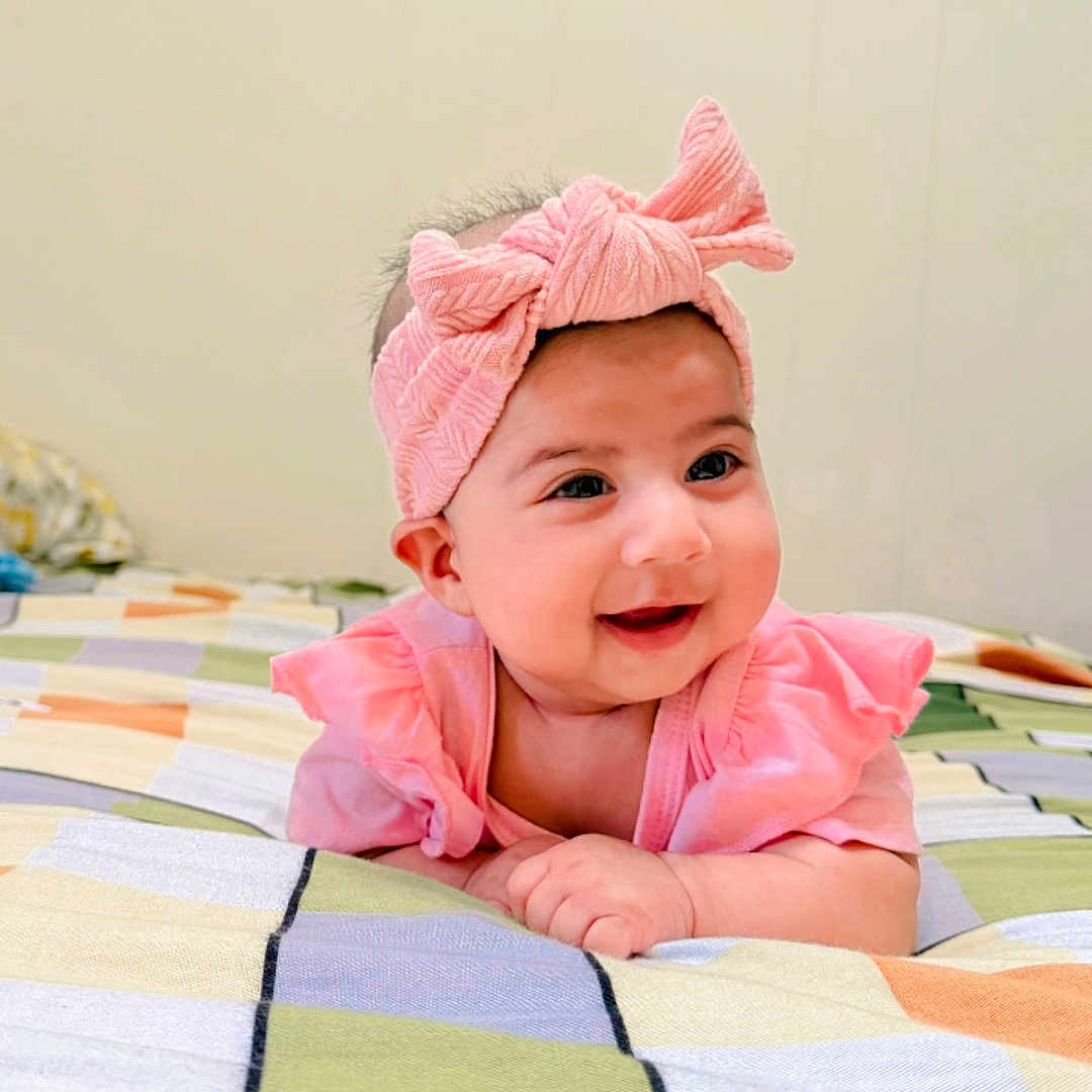 Doha Mehrish is registered to the contest to win money with this photo: baby, child, pink_bow, headband, smiling, tummy_pose, bedspread, colorful, checkered, indoor, cute, happy, infant, clothing, portrait, face, person, wall, soft_lighting, resting