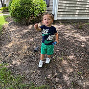 Zayden is registered to the contest to win money with this photo: toddler, child, outdoor, toy, nike, clothing, sneakers, grass, bush, house, sidewalk, curious, standing, daylight, person, young_child, shorts, tshirt, nature, play