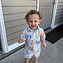 toddler, child, barefoot, curly_hair, smiling, outdoor, concrete, door, shirt, shorts, patterned_clothing, happy, standing, front_portrait, house_exterior, door_mat, sidewalk, summer, cute, person