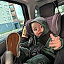 baby, car_interior, car_seat, casual_wear, child, daylight, face, footwear, green_clothing, hoodie, person, playful, seat, seatbelt, sneakers, toddler, transportation, urban_background, window, young_child