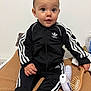 adidas, baby, black_clothing, blanket, bottle, child, clothing, curious, face, footwear, furniture, hairbrush, head, indoor, person, room, sitting, socks, tracksuit, young_child